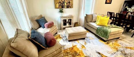 A living room with a sofa, ottoman, and a coffee table. There is a painting on the wall and a window with curtains.