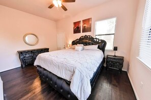 Quarto com cama grande, ventilador de teto, janela com persianas e espelho fixado na parede.