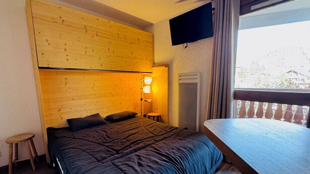Une chambre à coucher comprenant un lit, une table de chevet, une lampe, une fenêtre avec des rideaux et un téléviseur fixé au mur.
