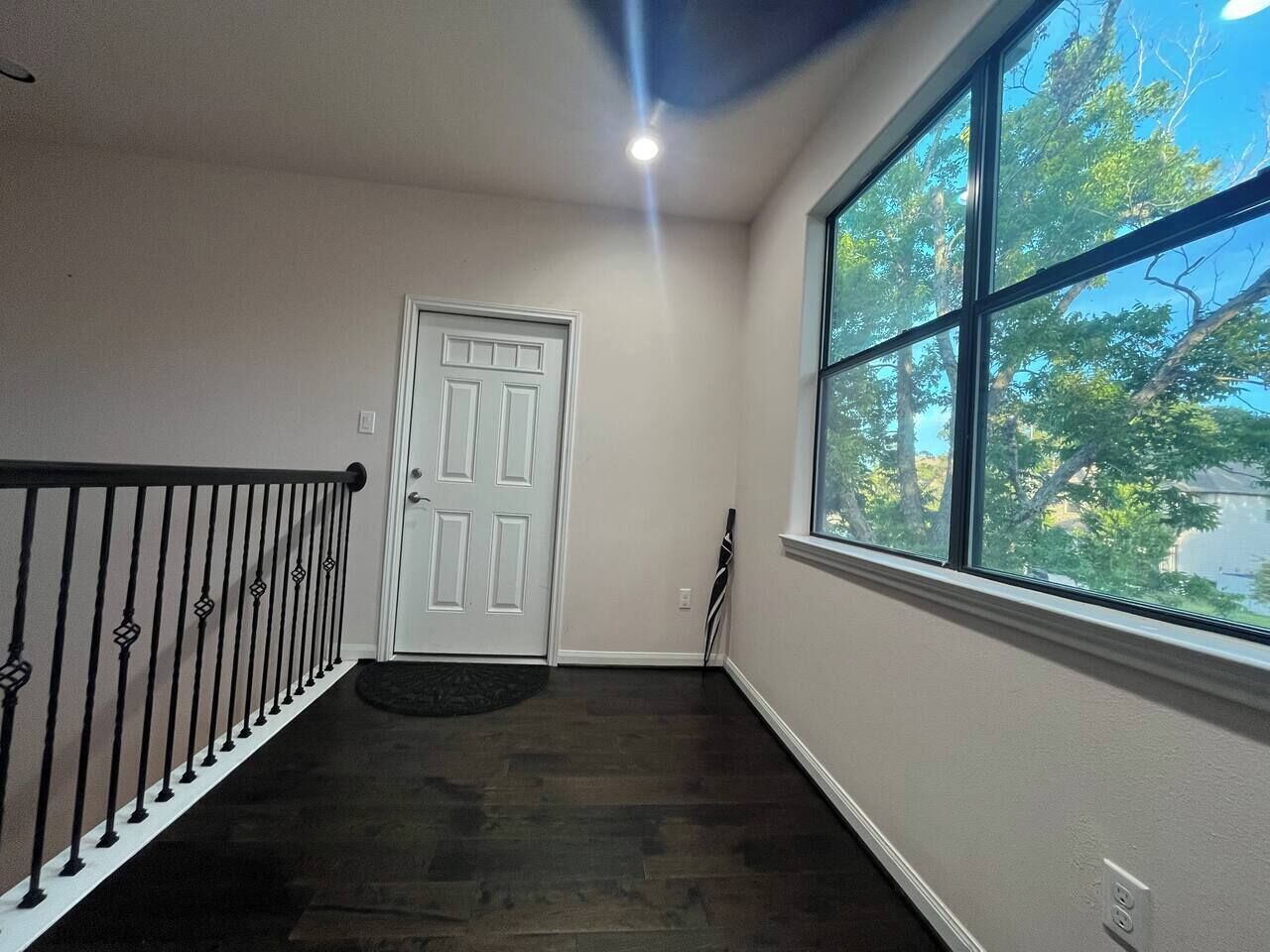 A hallway with a white door, a black railing, and a large window with a view of trees.