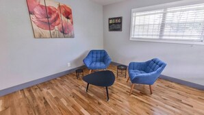 Ein Zimmer mit Holzboden, zwei blaue Sessel, ein schwarzer Hocker, ein kleiner runder Tisch und ein Gemälde mit roten Blumen an der Wand.