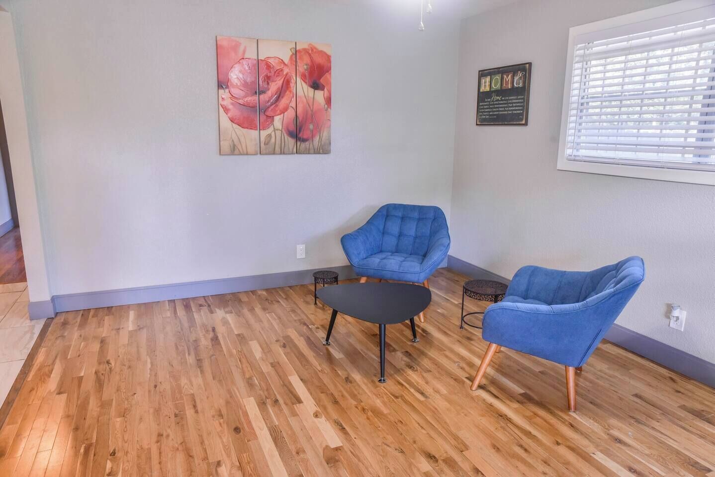 A room with wooden flooring, two blue armchairs, a small black table, a painting of red flowers, and a window with blinds.