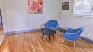 Ein Zimmer mit Holzboden, zwei blaue Sessel, ein kleiner schwarzer Tisch, ein Gemälde mit roten Blumen und ein Fenster mit Jalousien.