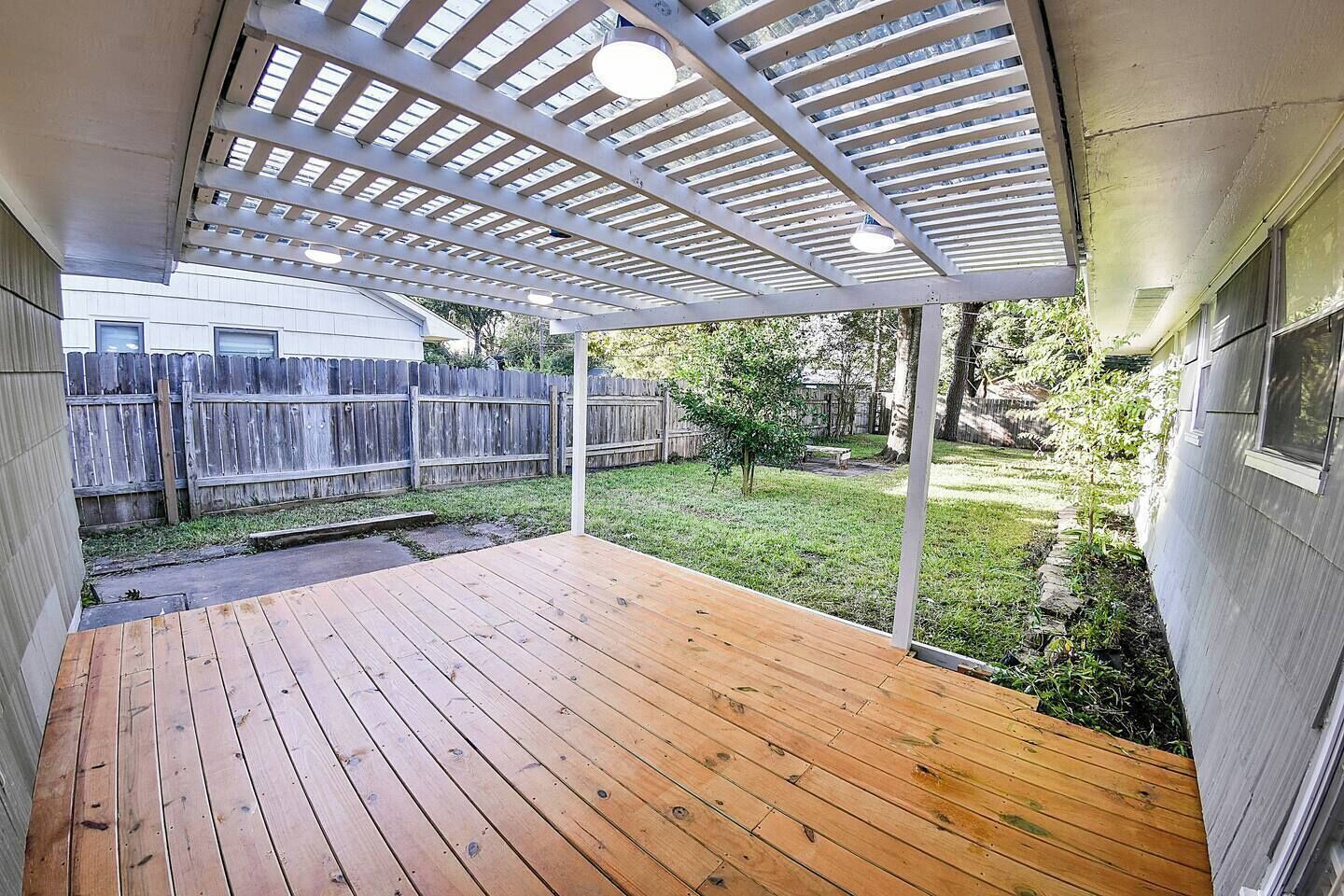 A wooden deck with a pergola overhead, a grassy yard, and a wooden fence in the background.