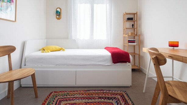 Ein Schlafzimmer mit einem Bett, einem Schreibtisch, einem Stuhl, einem Bücherregal und einem farbenfrohen Teppich.