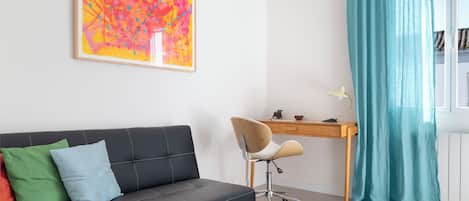 A modern living room with a black sofa, colorful cushions, a wooden desk, a chair, and a large abstract painting on the wall.