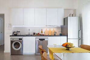 A modern kitchen with a washing machine, dishwasher, and a dining area with a table set for two. - Oasis Seville – Light-Filled Apartment with Terrace in Los Remedios (Sevilla)