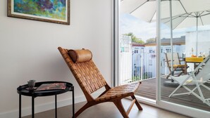 Ein Korbstuhl mit Kissen, ein kleiner Tisch mit Tasse und Buch sowie ein großes Fenster mit Blick auf einen Balkon und Gartenmöbel.
