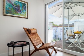 A wicker chair with a cushion, a small table with a cup and a book, and a large window with a view of a balcony and outdoor furniture. - Oasis Seville – Light-Filled Apartment with Terrace in Los Remedios (Sevilla)