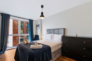 Quarto de hotel com cama, penteadeira, cadeira e janela com cortinas.