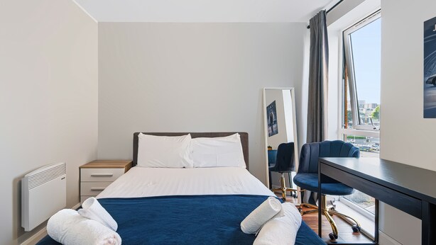 A hotel room with a bed, a desk, a chair, a radiator, and a window with a view of the city. - Stunning 2BDR 2Bath Salford Apartment with Parking (Greater Manchester)