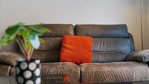 A leather sofa with a single orange cushion, a glass coffee table, and a potted plant in the foreground. - Stunning 2BDR 2Bath Salford Apartment with Parking (Greater Manchester)