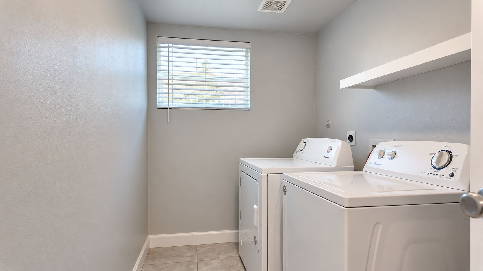 A laundry room with a washer and dryer, a window with blinds, and a white shelf.