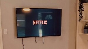 Een flatscreen televisie bevestigd aan een muur, met het Netflix logo, een witte console tafel eronder en een houten vloer.
