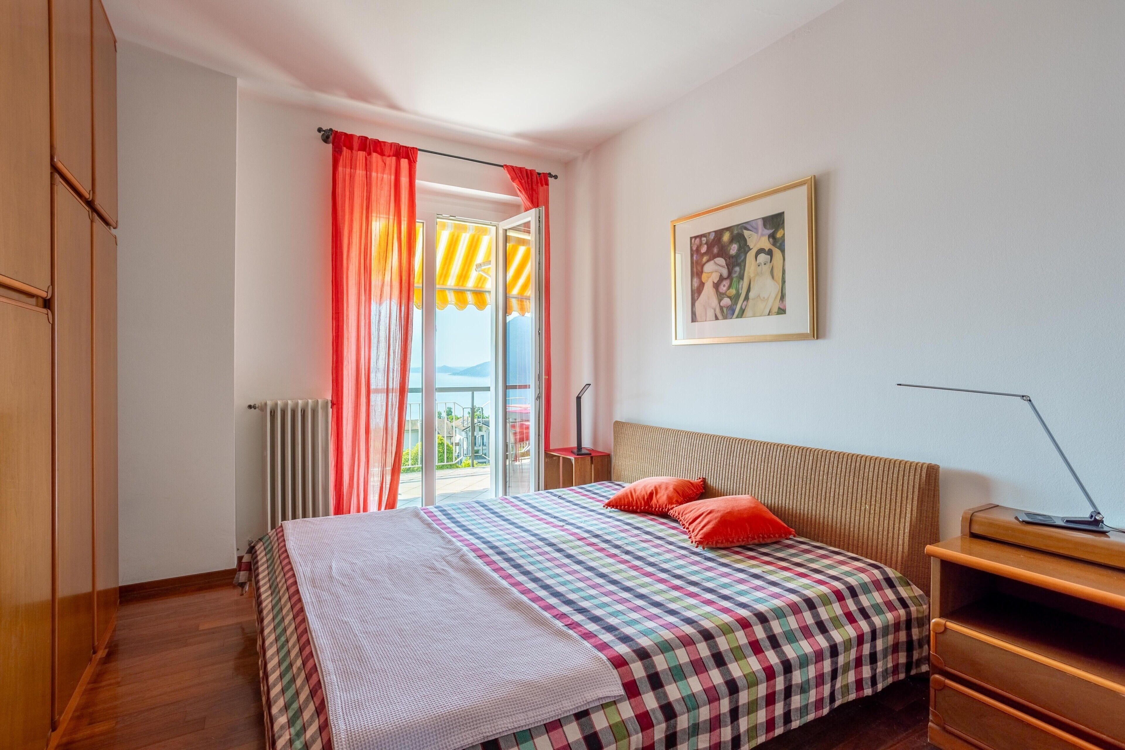 Un dormitorio con cama, mesita de noche, lámpara, radiador, ventana con cortinas y un cuadro en la pared.