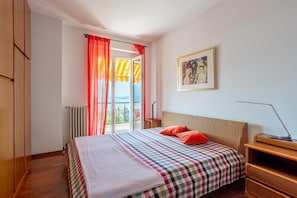 Un dormitorio con cama, mesita de noche, lámpara, radiador, ventana con cortinas y un cuadro en la pared.