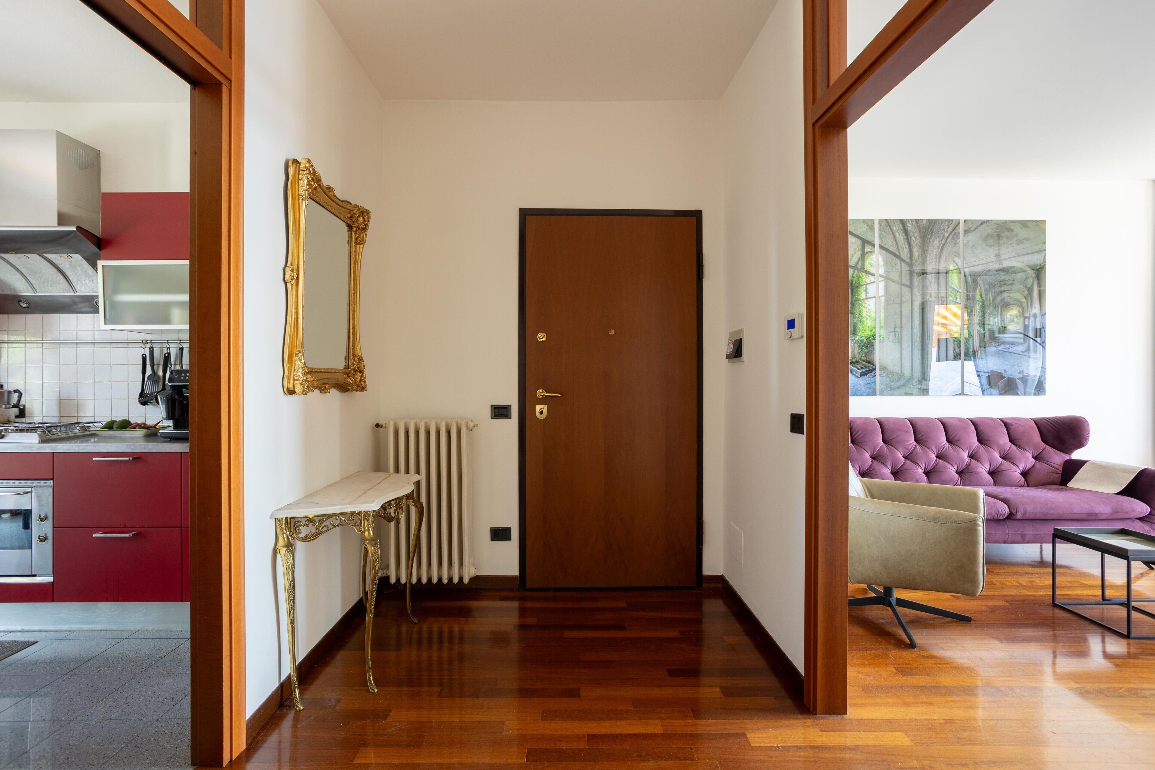Un corridoio con pavimento in legno, una porta, un termosifone, un tavolo con cornice dorata e un divano viola.
