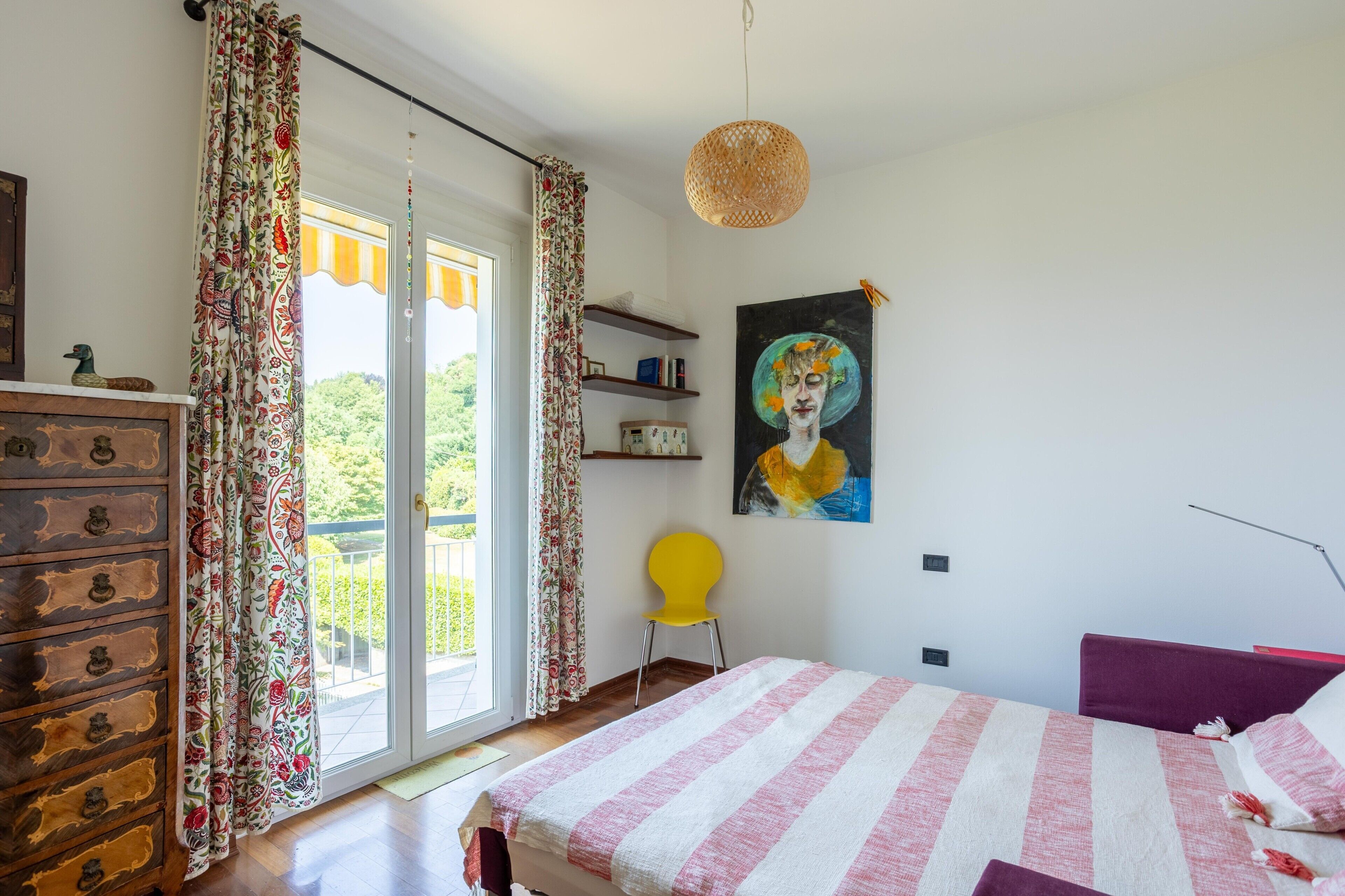 Quarto com cama, cômoda, cadeira, pintura e janela com cortinas.