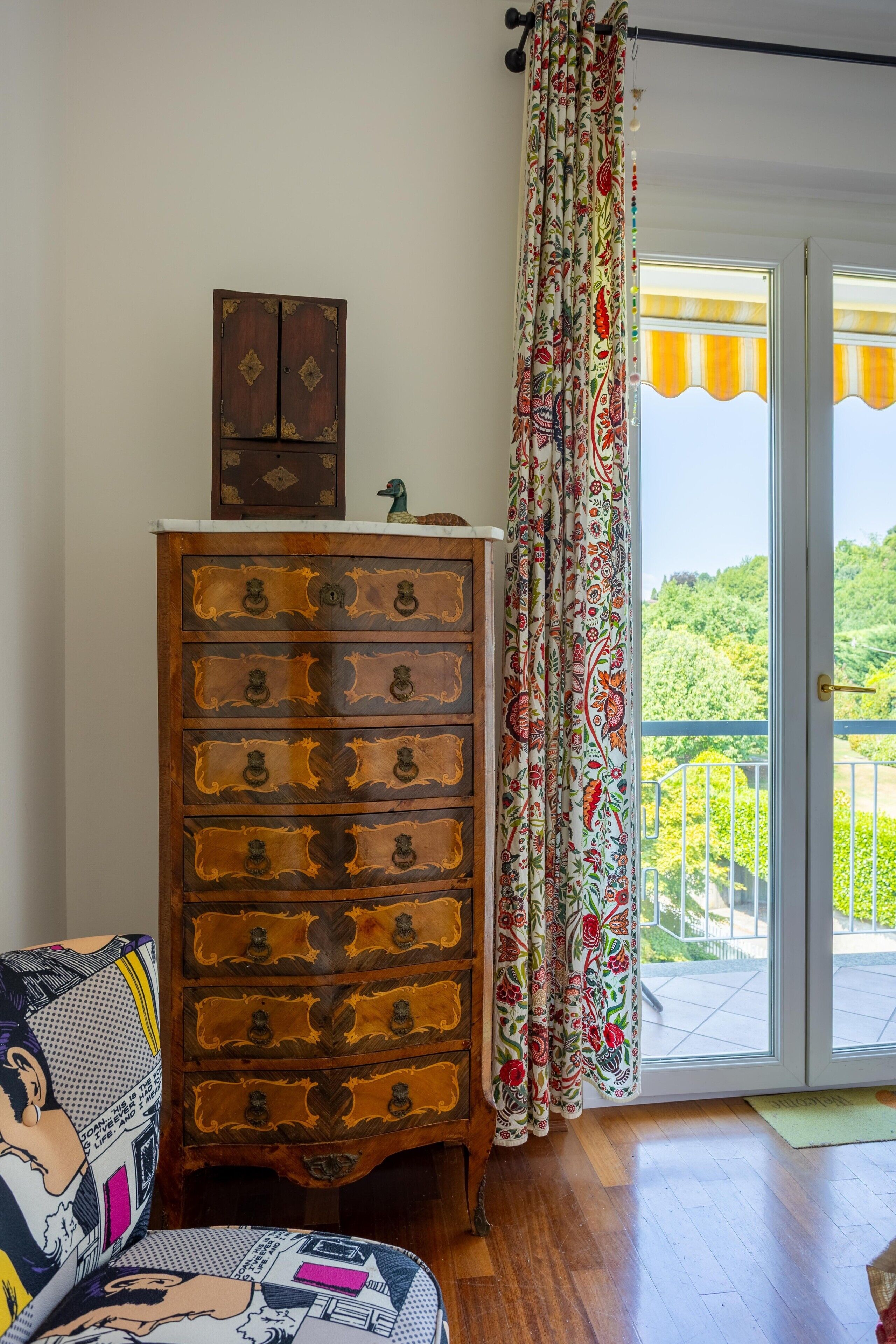 Un mobile contenitore in legno con maniglie decorate, una statuina di un'anatra ornamentale e una tenda colorata con motivi floreali.