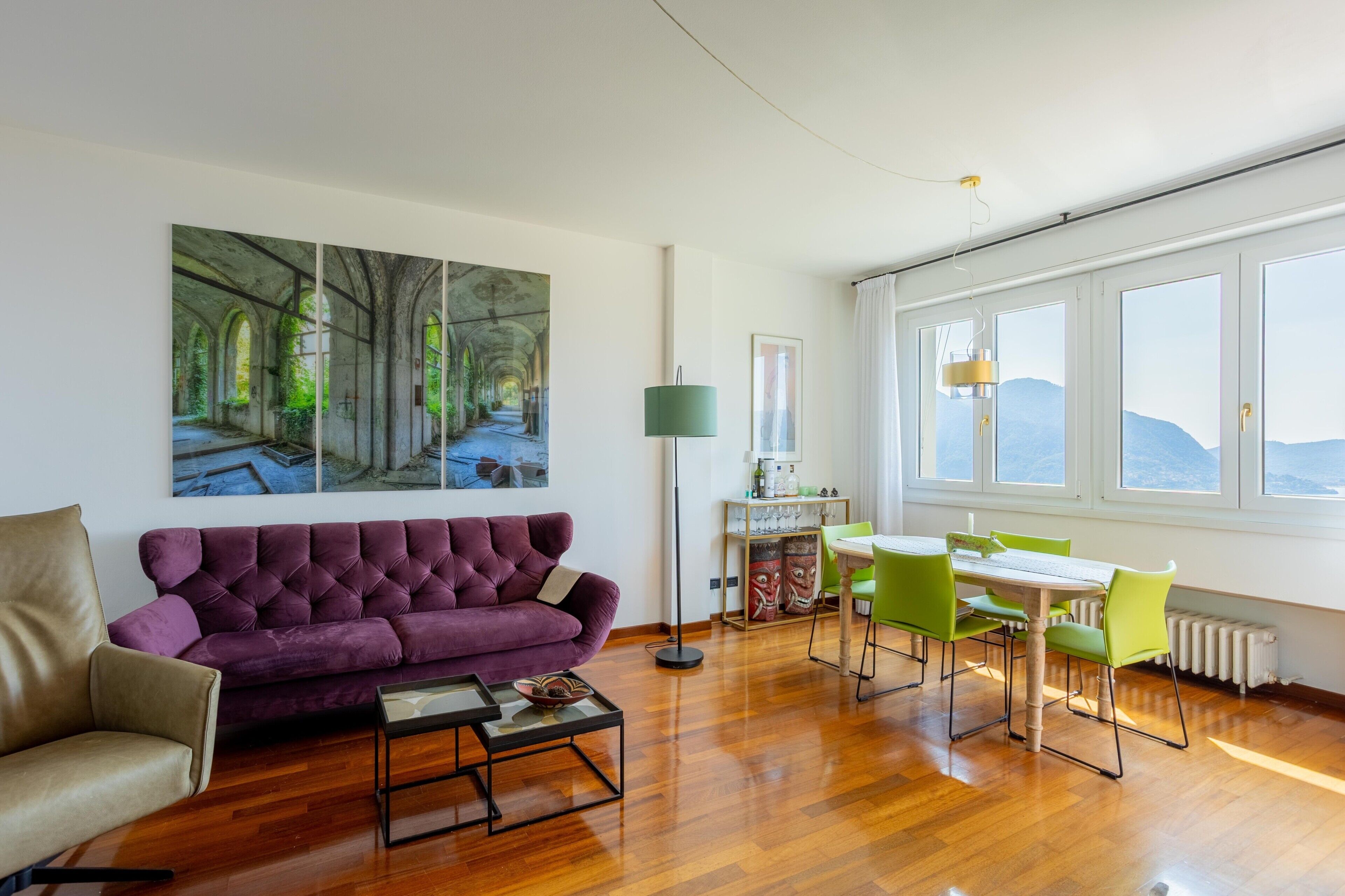 Een kamer met een paarse bank, een eettafel met groene stoelen en een groot raam met uitzicht.