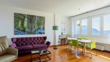 Een kamer met een paarse bank, een eettafel met groene stoelen en een groot raam met uitzicht.