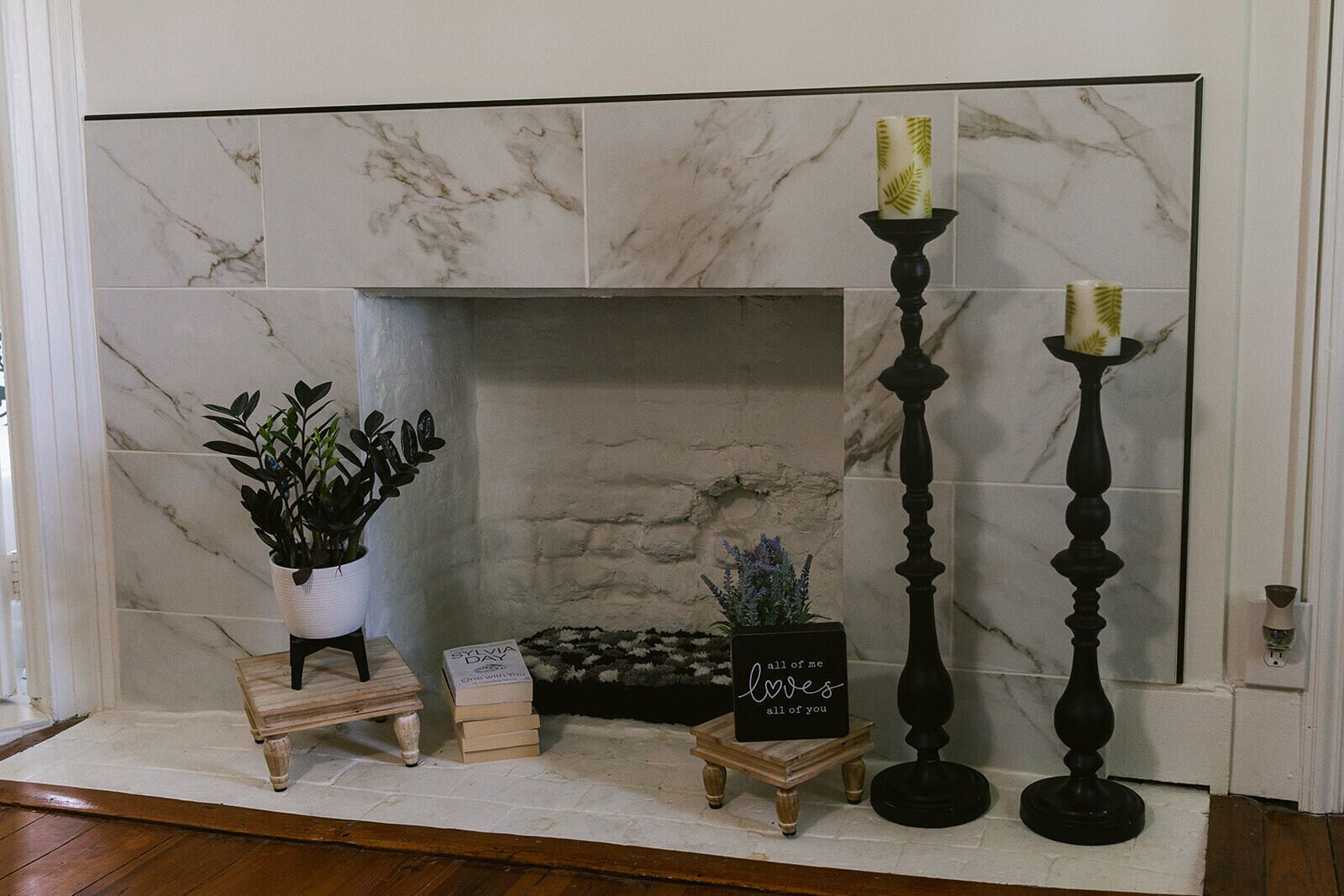 Une cheminée avec un manteau en pierre, une plante en pot, une petite table en bois sur laquelle sont posés des livres, et deux hauts chandeliers.