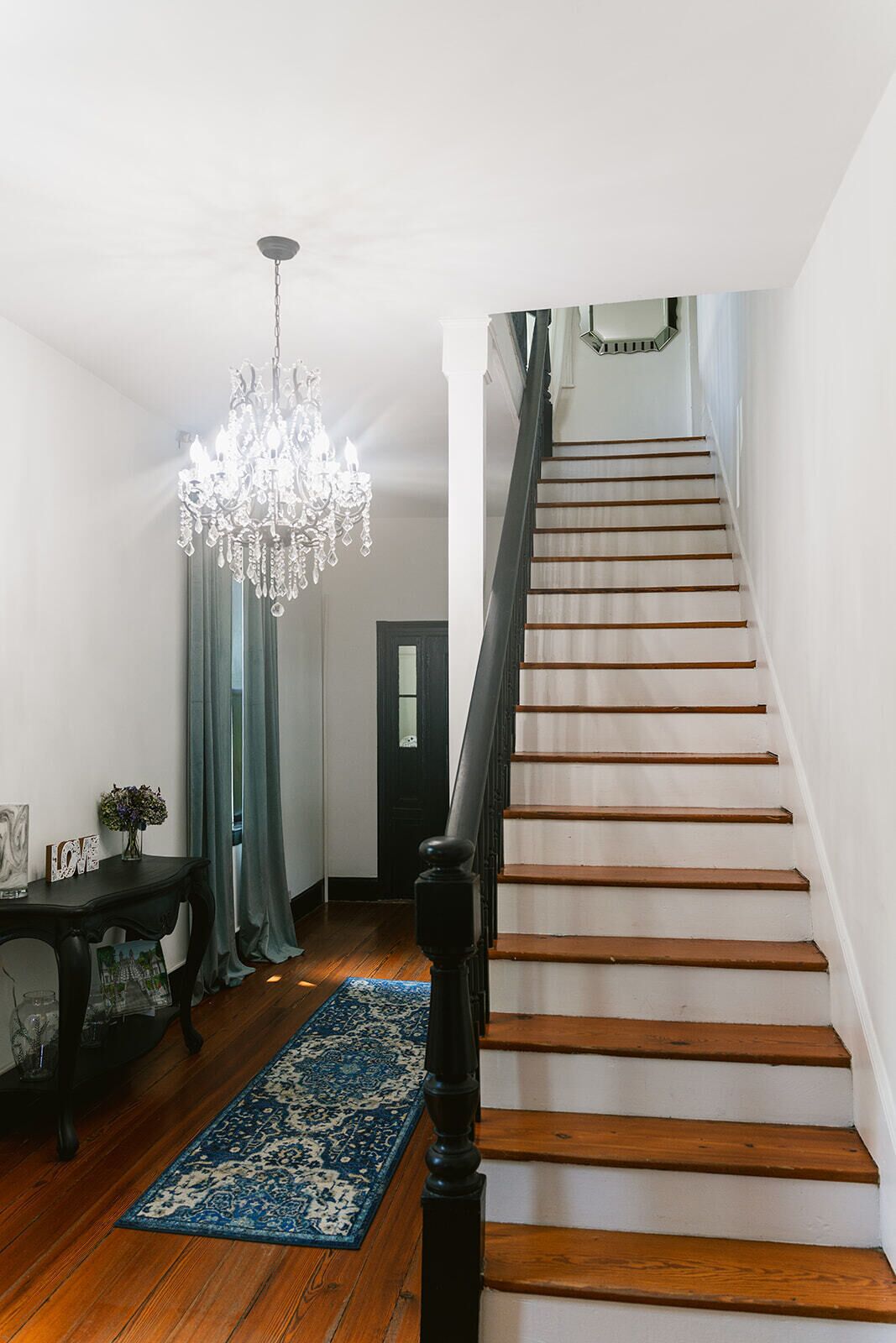 A staircase with wooden steps and a black handrail, leading to a landing with a chandelier and a table with a rug.