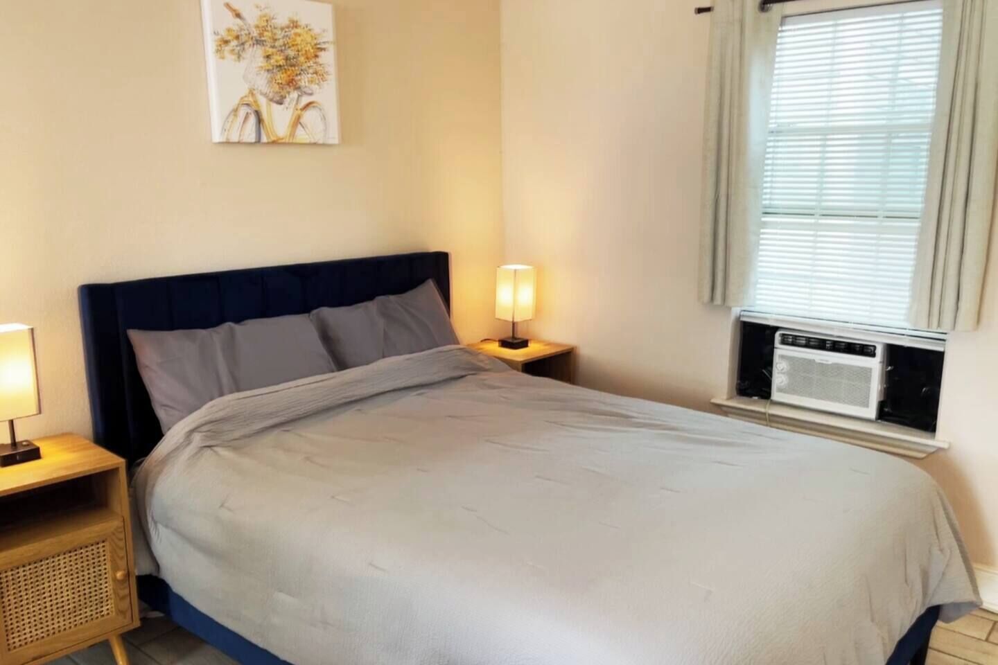 A bed with a dark headboard, two bedside lamps, a wall-mounted air conditioning unit, and a window with blinds.