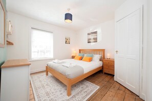 A bedroom with a wooden bed, a nightstand, a lamp, a window with blinds, and a door. - Charming Cottage 15 minutes to central London - Pass the Keys (Surbiton)