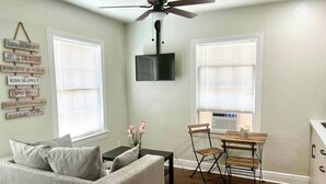 A living room with a gray sofa, a coffee table, a TV mounted on the wall, a ceiling fan, and a dining area with a table and chairs. - Cozy Retreat w/Coffee Bar & Private Entry (Houston)
