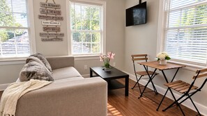 Ein Wohnzimmer mit einer Couch, einem kleinen Tisch mit Blumen und einem an der Wand montierten Fernseher.