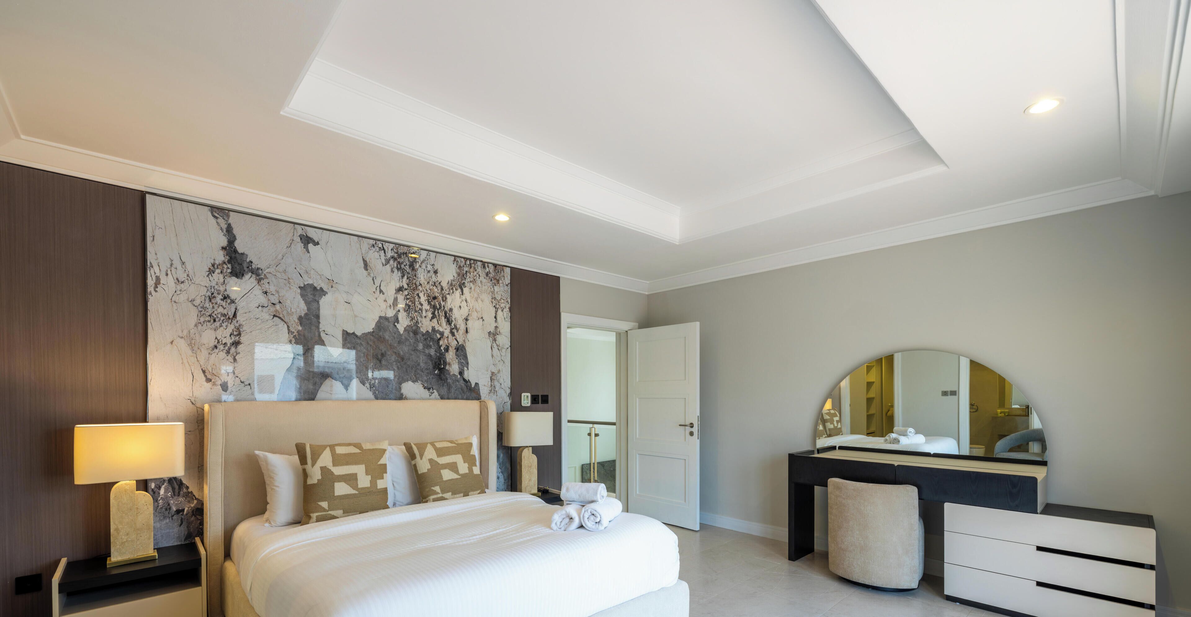 A hotel room with a bed, bedside tables, a dresser, a mirror, and a large abstract wall art piece.