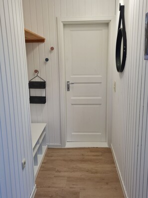 Een smalle gang met een witte deur, houten vloer en een zwarte ophanghaak aan de muur.