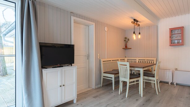 A room with a television on a cabinet, a bench, a table with chairs, and a wall-mounted light fixture. - Modern Eingerichteter Doppelbungalow mit Garten in Mirow (Mirow)