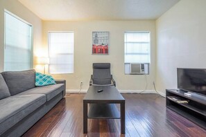 A living room with a sofa, a coffee table, a TV, and two windows with blinds. - Pet-Friendly Houston Apartment w\ Backyard (Houston)