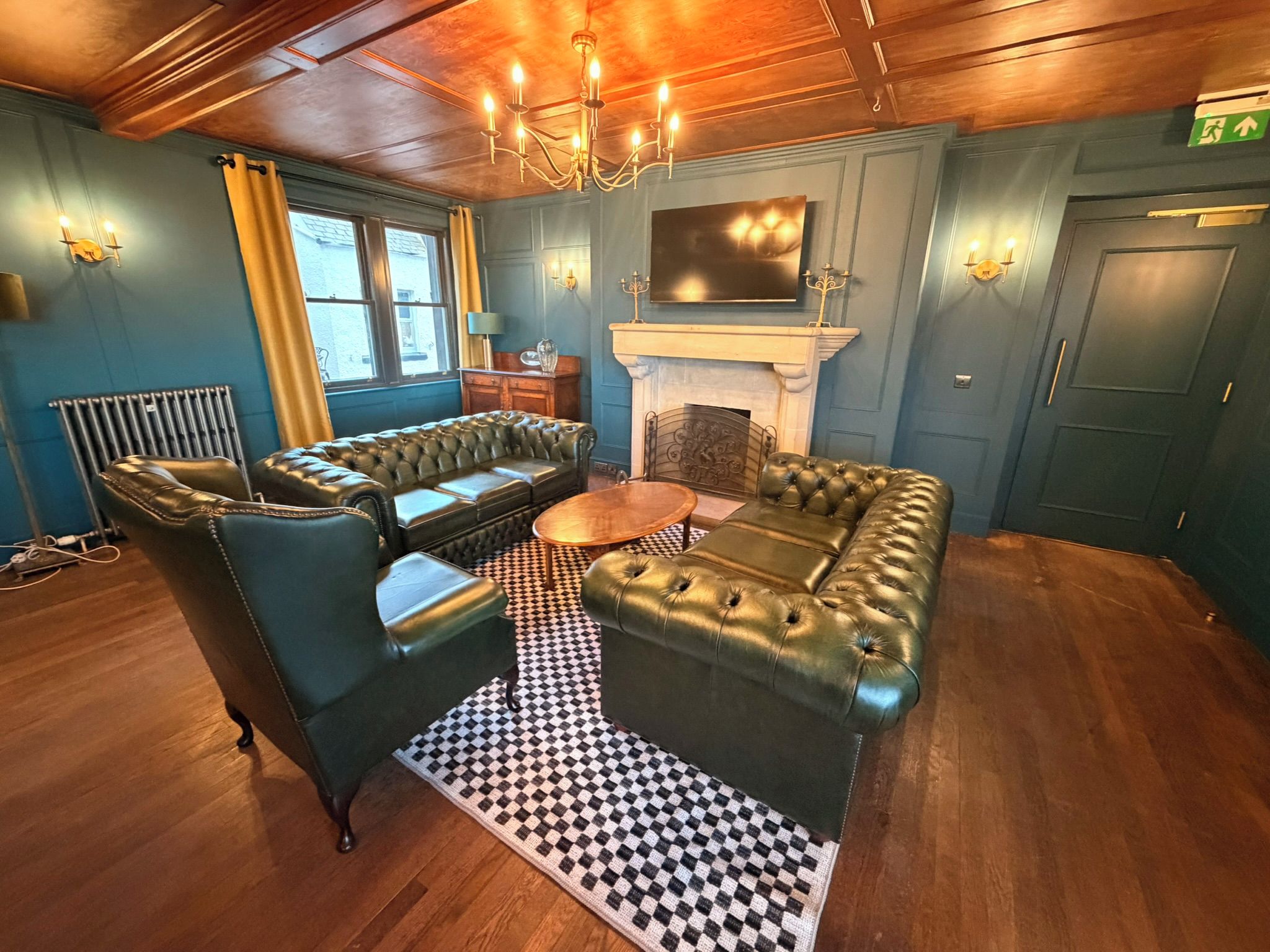 A room with a checkered floor, a fireplace, a television mounted above it, and a chandelier.