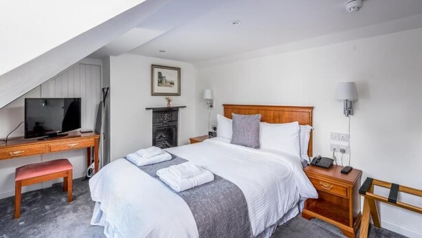 A hotel room with a bed, a desk with a computer, a chair, a bedside table with a lamp, and a fireplace.