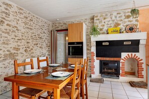 A dining area with a wooden table and chairs, a kitchen with a built-in oven, and a brick fireplace with a clock above it. - Bournezeau Holiday Cottage in the Countryside, Near Coast and Attractions (Bournezeau)