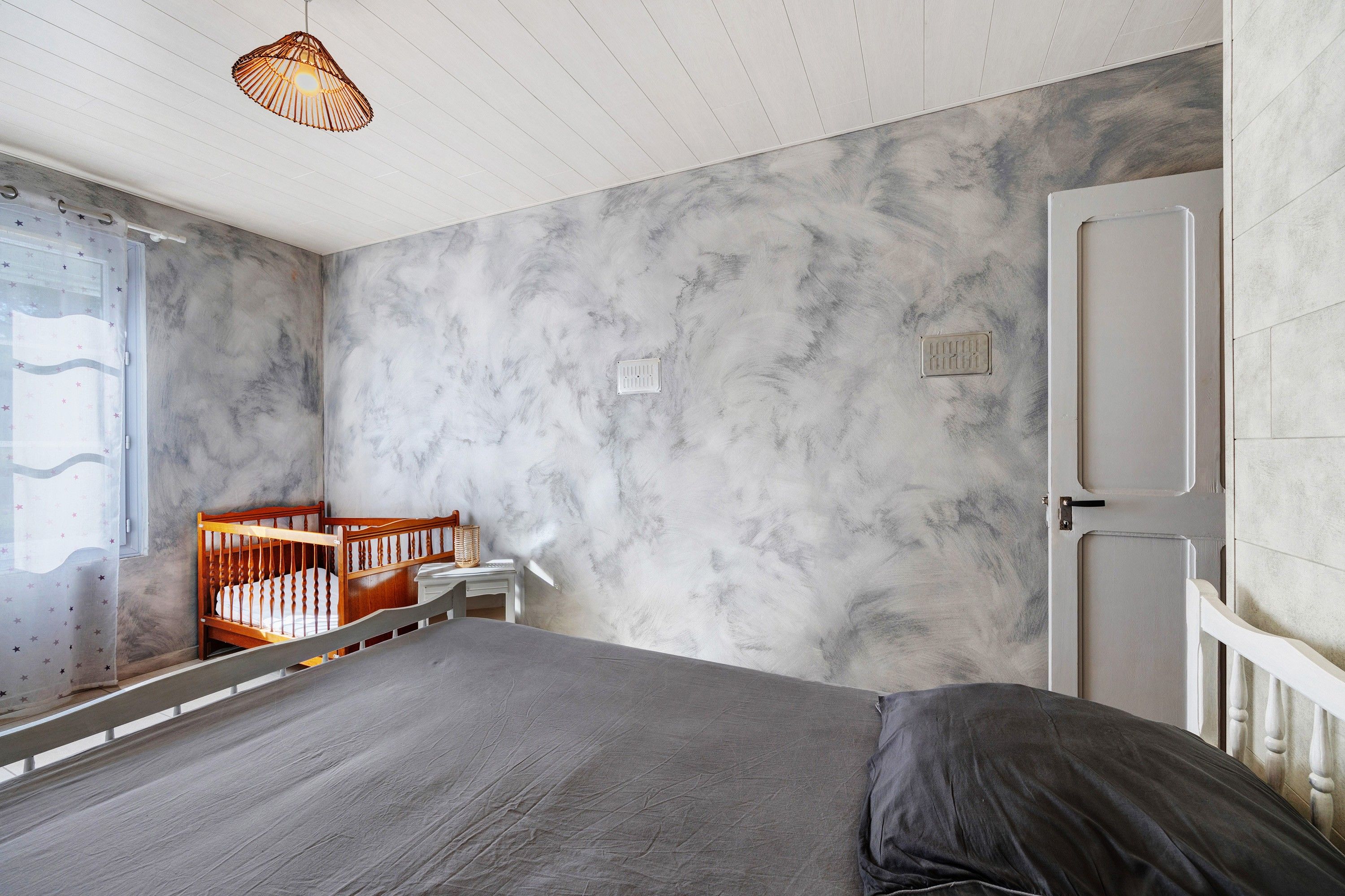 Une chambre à coucher avec un lit, un berceau, une fenêtre ornée d’un rideau à motifs d’étoiles et un mur à texture.