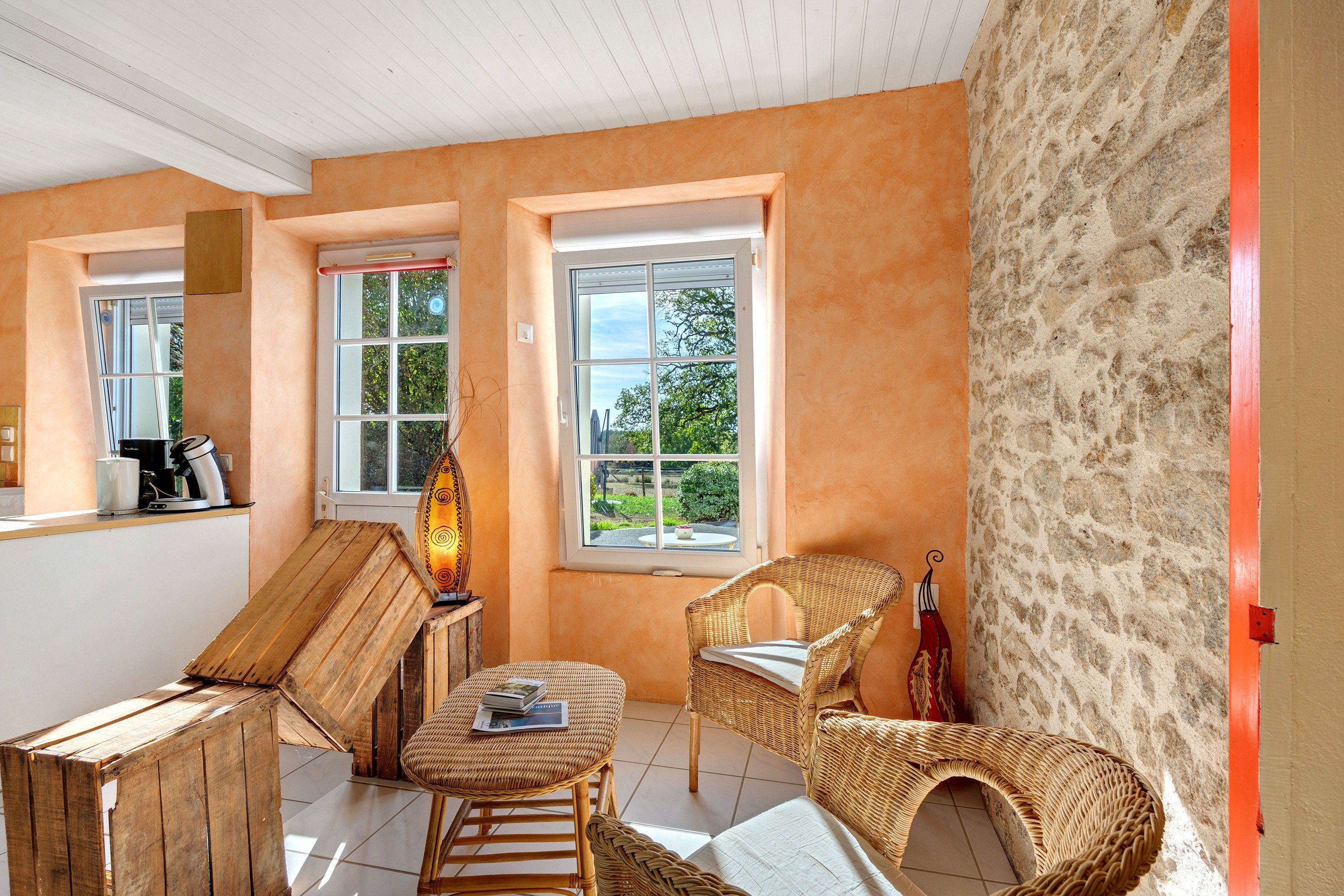 Une pièce chaleureuse avec un banc en bois, deux fauteuils en osier, une table avec une lampe et une vue sur la verdure par les fenêtres.