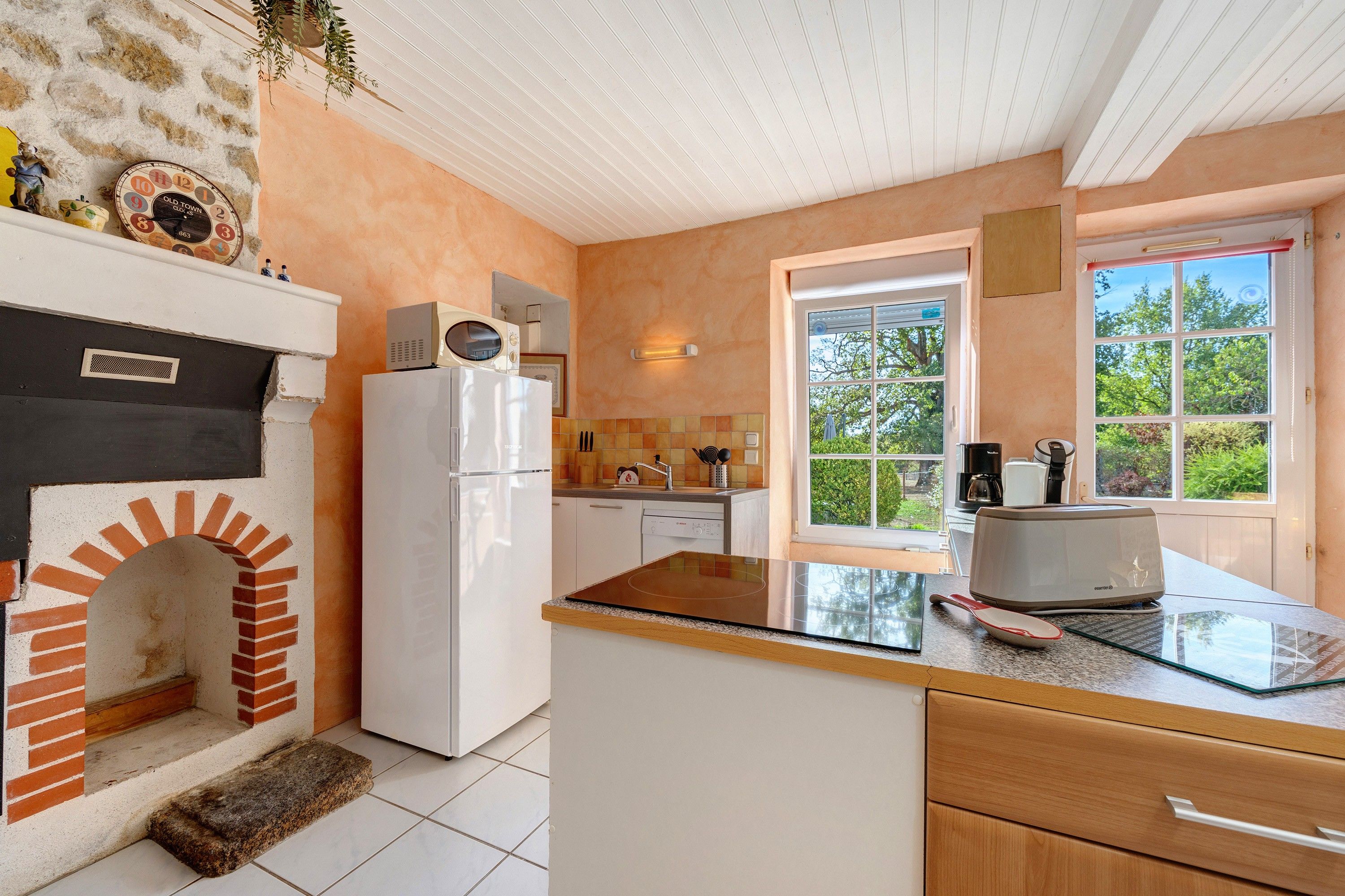 A kitchen with a brick fireplace, a white refrigerator, and a window with a view of greenery.