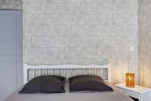 Ein Bett mit weißem Kopfteil, zwei dunklen Kissen und einem Nachttisch mit Lampe.