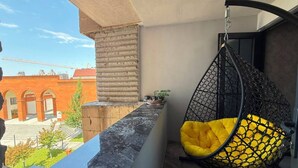 Un balcón con una silla colgante, vista a un edificio rojo y un muro de piedra.
