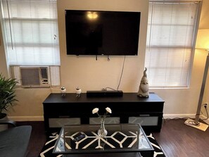 A living room with a flat-screen TV mounted on the wall, a black entertainment unit, a glass coffee table with a geometric pattern, and a vase with flowers. - Pet-Friendly Home w/ Patio & Backyard (Houston)