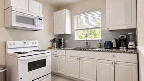 A kitchen with white cabinets, a stove, a microwave, a window with blinds, and various kitchen appliances on the countertop. - Pet-Friendly Home w/ Patio & Backyard (Houston)