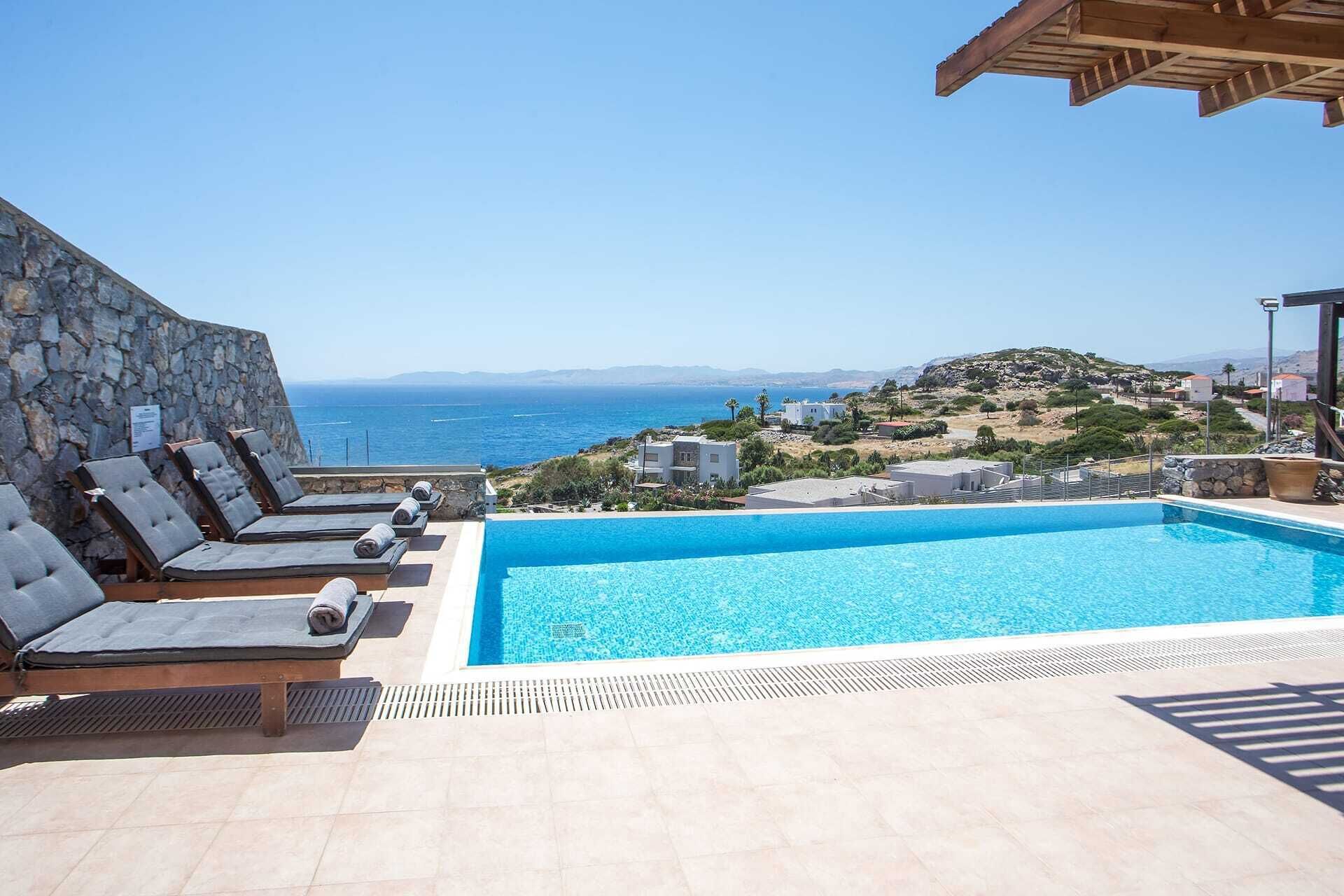 En pool med solstolar, en stenmur och utsikt över havet och kullarna.