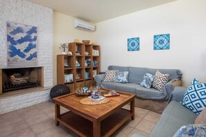 A living room with a fireplace, a wooden coffee table, a sofa with patterned cushions, a bookshelf, and two wall-mounted decorative plates. - Miranda Infinity Pool Villa Pefkos Rhodes (Rhodes)