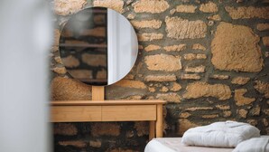 Una habitación con pared de piedra, un espejo redondo, una mesita de noche de madera y una cama con manta blanca.