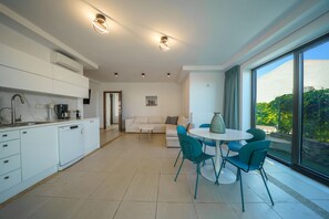 A modern kitchen with white cabinets, a dining area with a round table and teal chairs, and a large window offering a view of greenery. - Crethion | Private Pool, Jacuzzi, Gym & Games Room (Violi Charaki)