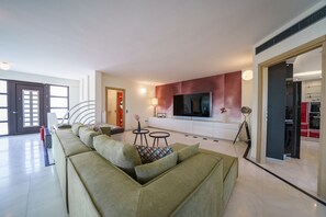 A modern living room with a sectional sofa, a flat-screen TV mounted on the wall, and a small dining area with a table and chairs. - Crethion | Private Pool, Jacuzzi, Gym & Games Room (Violi Charaki)